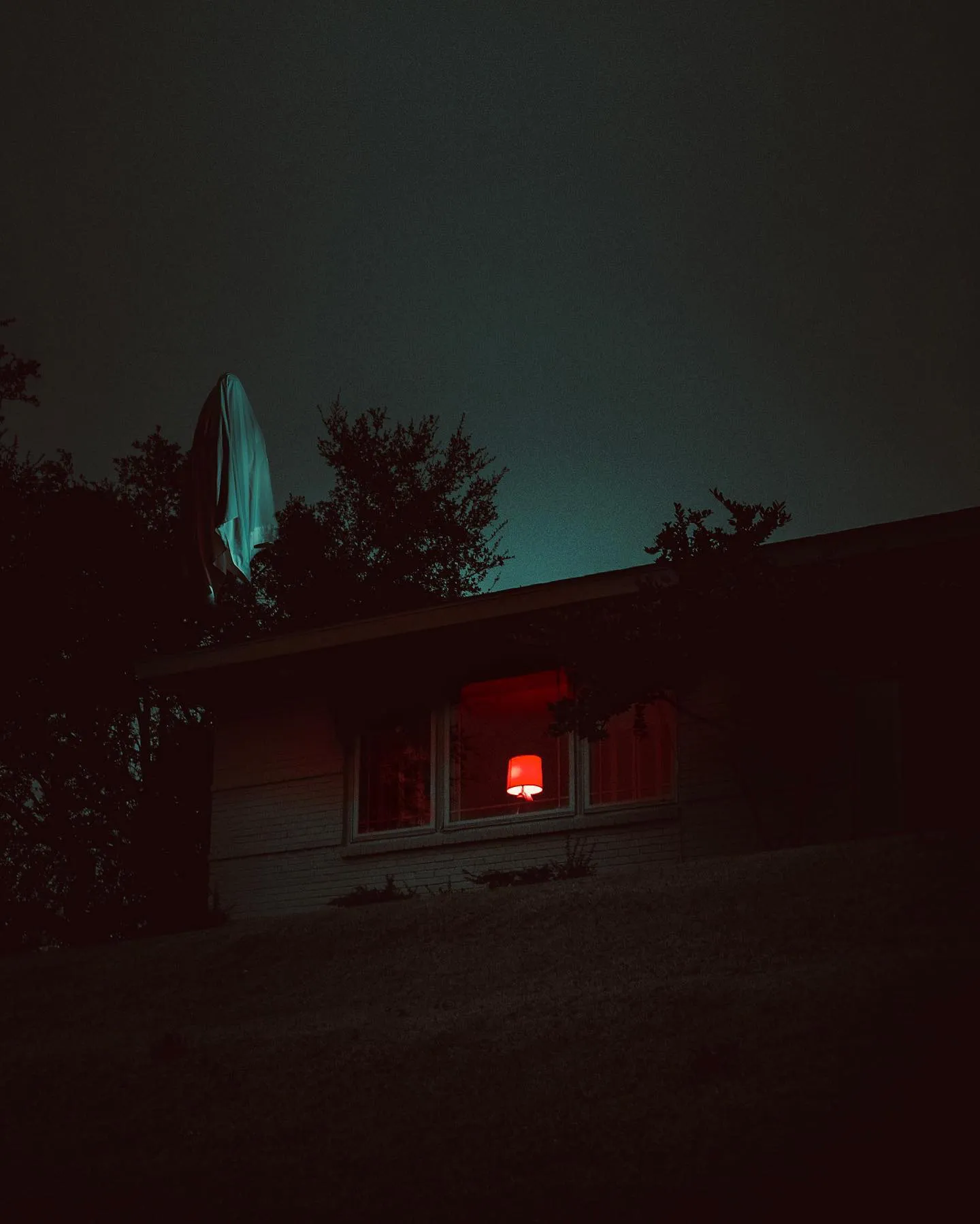 A dark house at night; one window glows red, a white cloth form hangs in the yard.
