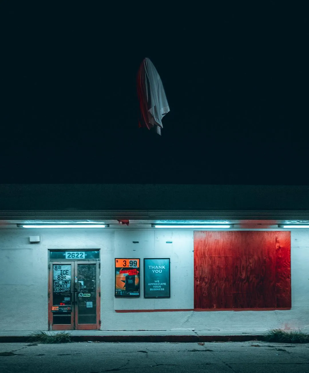A storefront at night with a red door panel and white light, a white cloth figure floating above the roofline.