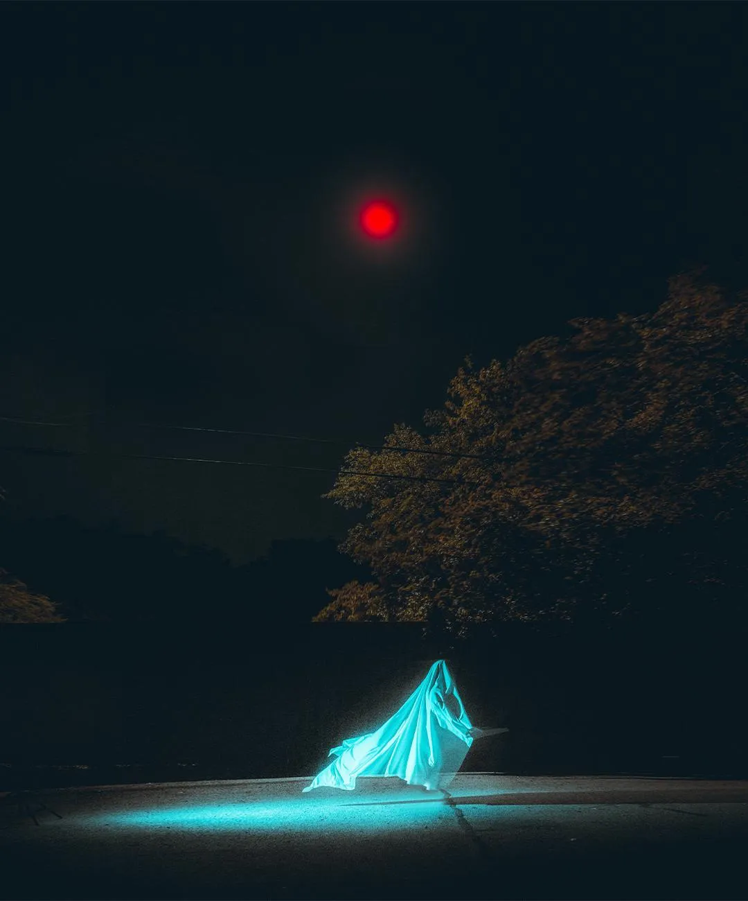 A teal-lit draped cloth figure in a park at night, a red street lamp overhead.