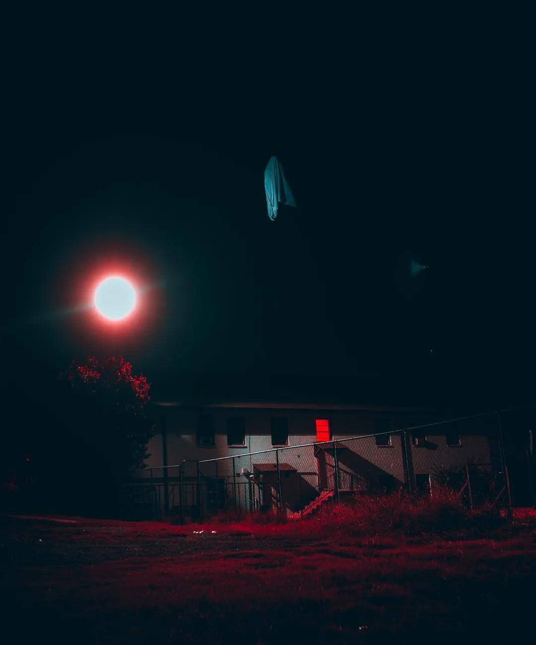 A red circular light above a draped cloth figure, a house behind it in blue night.