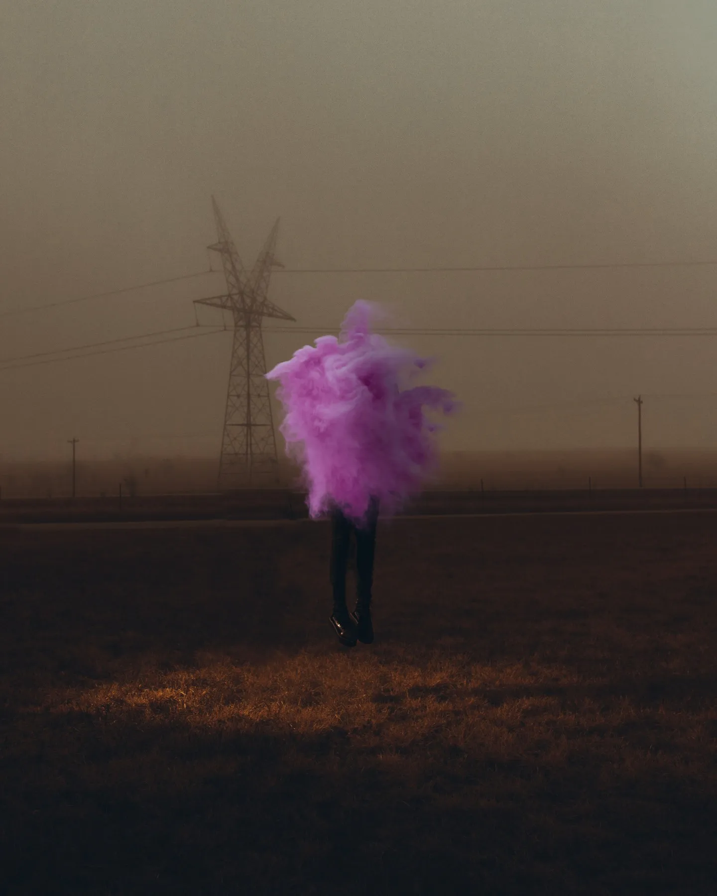A figure in a pink dress standing in a foggy field with a transmission tower behind, muted colour.