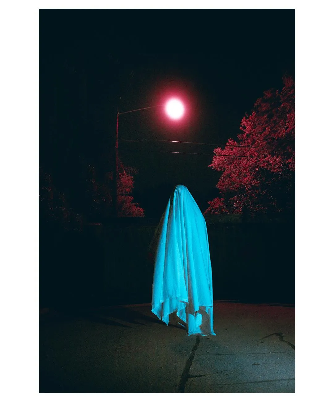 A blue-lit draped cloth figure beneath a street lamp at night.