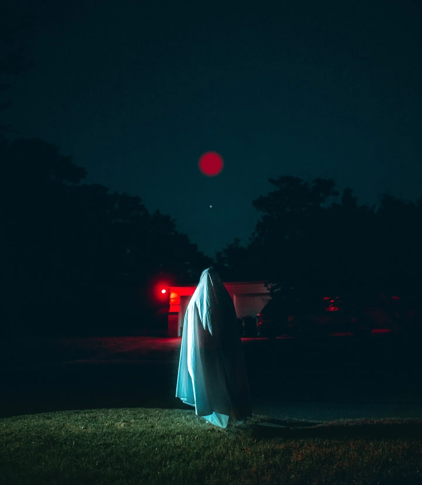 A figure in white cloth standing in a park at dusk, a red light nearby.