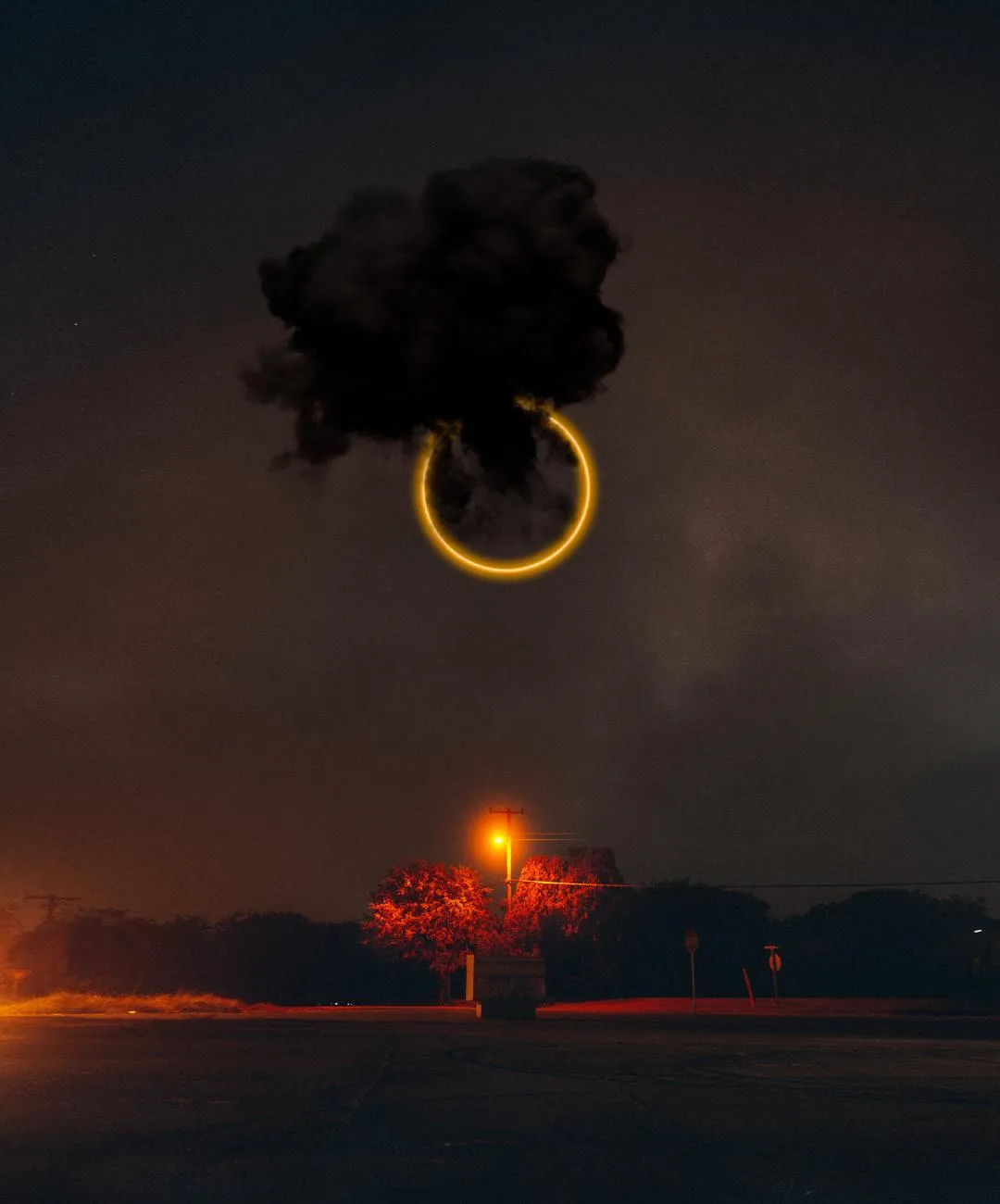 A black cloud of smoke with a pale gold ring above it over a dusk horizon and an orange streetlight.